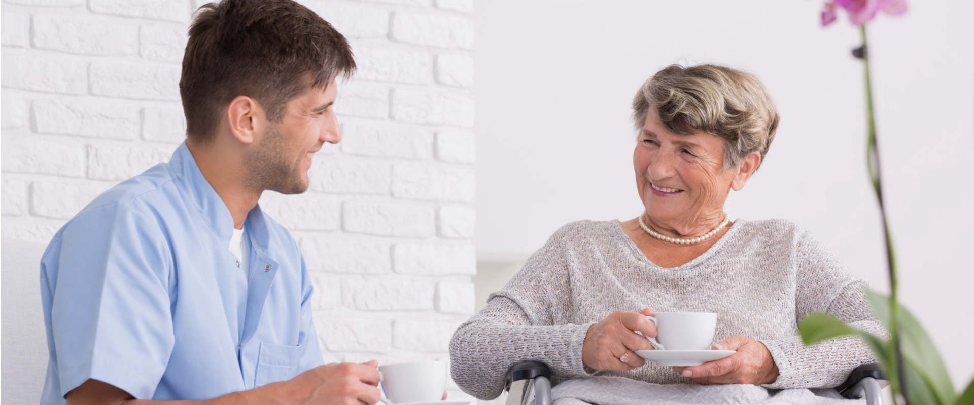 A brunette young white man speaking with an geriatric woman in a wheel chair while both are sipping tea.